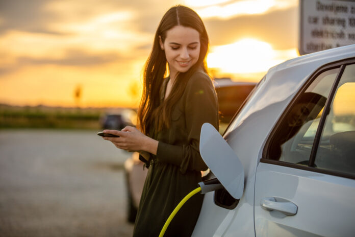 Woman waiting for electric car to charge in the parking lot at sunset